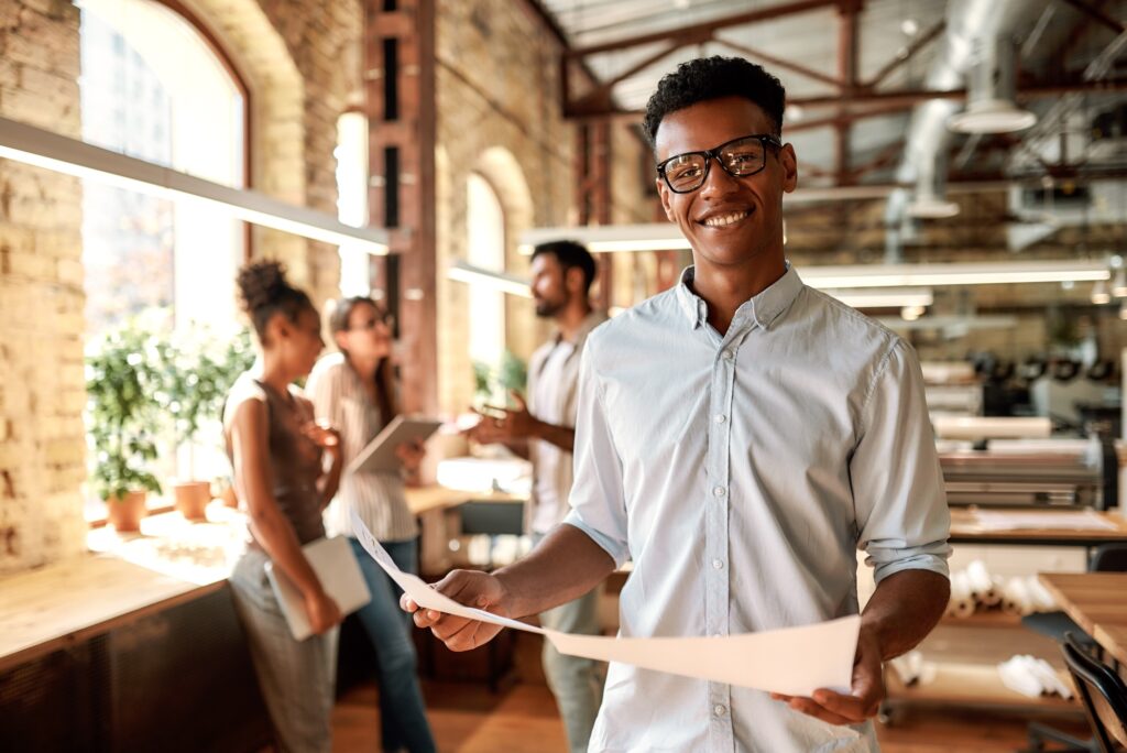 Enjoying my work. Young afro american man holding documents and looking at camera with smile while